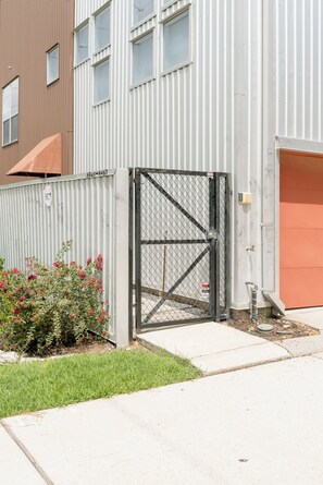 A gated entrance to a building with a metal fence, a small garden with pink flowers, and a concrete pathway leading to a garage. - Urban Luxe Escape - Stylish & Serene (Houston)