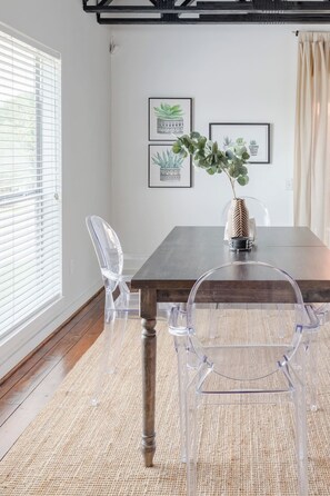 A dining area with a wooden table, clear plastic chairs, a woven rug, and framed plant illustrations on the wall. - Urban Luxe Escape - Stylish & Serene (Houston)