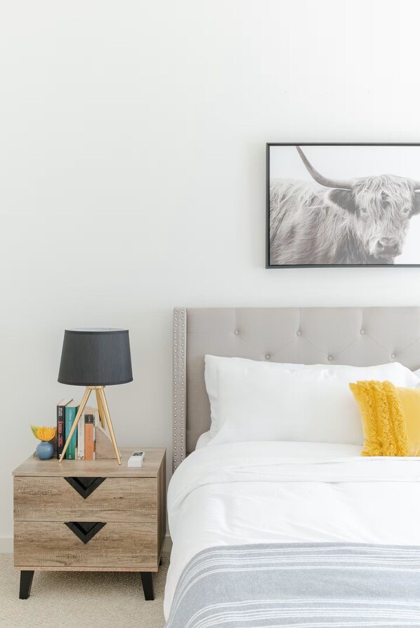 A bedroom with a bed, a bedside table with a lamp, a framed picture on the wall, and a nightstand with books and a vase. - Urban Luxe Escape - Stylish & Serene (Houston)