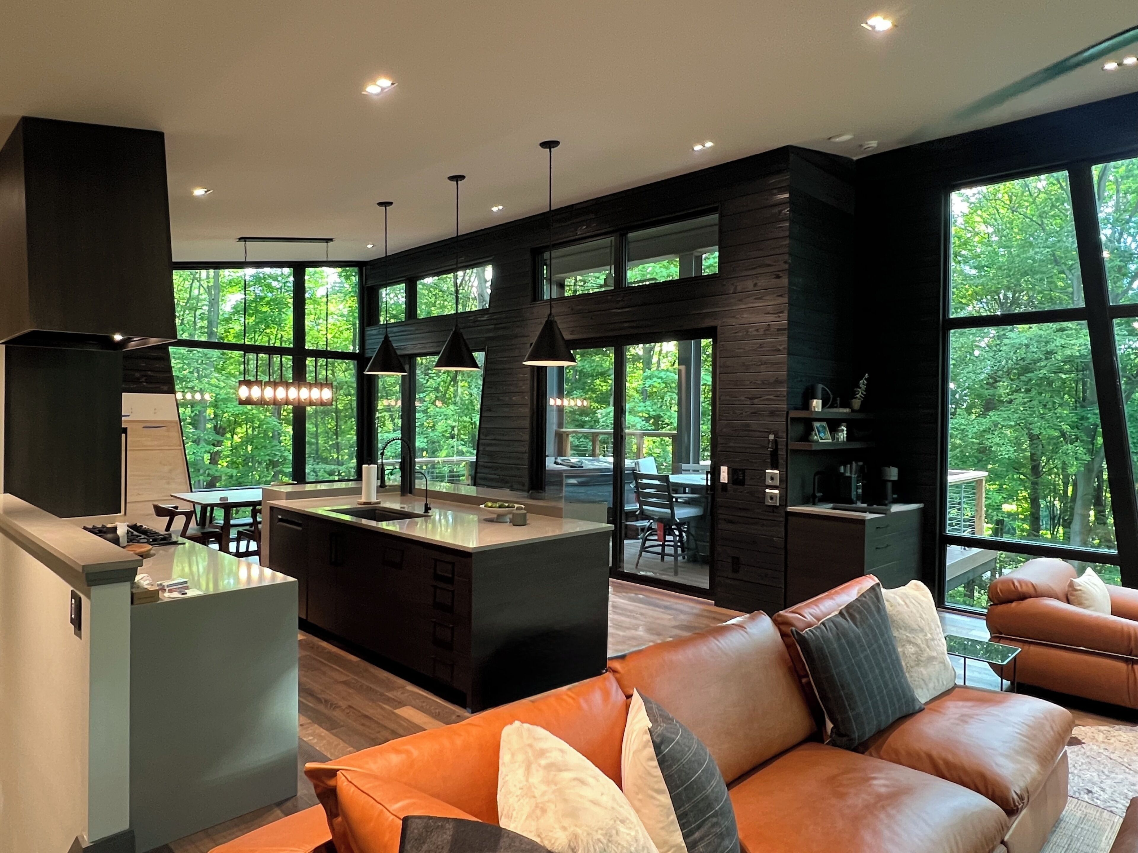 A modern kitchen with a central island, dining area, and large windows offering a view of the outdoors.