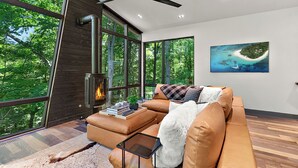 A modern living room with a large window, a fireplace, a sofa, and a painting of a tropical island. - The Treehouse at Norstead (Cedar)