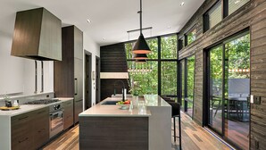 A modern kitchen with a central island, stainless steel appliances, and a large window providing a view of the outdoors. - The Treehouse at Norstead (Cedar)