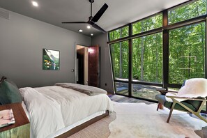 Ein Schlafzimmer mit einem großen Bett, einem Deckenventilator, Blick auf einen Wald durch große Fenster und einer Tür zu einem anderen Raum.