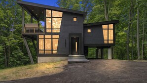 A modern two-story house with large windows and a balcony, surrounded by a forest. - The Treehouse at Norstead (Cedar)