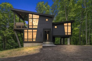 A modern two-story house with large windows and a balcony, surrounded by a forest.