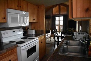 A kitchen with wooden cabinets, a white stove, a microwave, and a dining area with a table and chairs. - Acadian Shores Cabins - Lake View Cabin 4 (Zwolle)