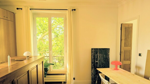 A kitchen with wooden cabinets, a window with curtains, a dining table with chairs, and a door. - Parisian charm, sunny apartment, beautiful views (Paris)