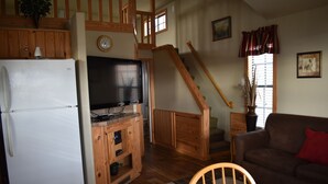 A kitchen with a refrigerator, a wooden cabinet, and a television. There is a staircase leading to an upper level.