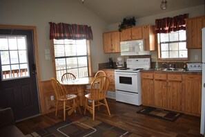 A compact kitchen with wooden cabinets, a stove, a microwave, and a dining area with chairs and a table. - Acadian Shores Cabins - Lake View Cabin 2 (Zwolle)