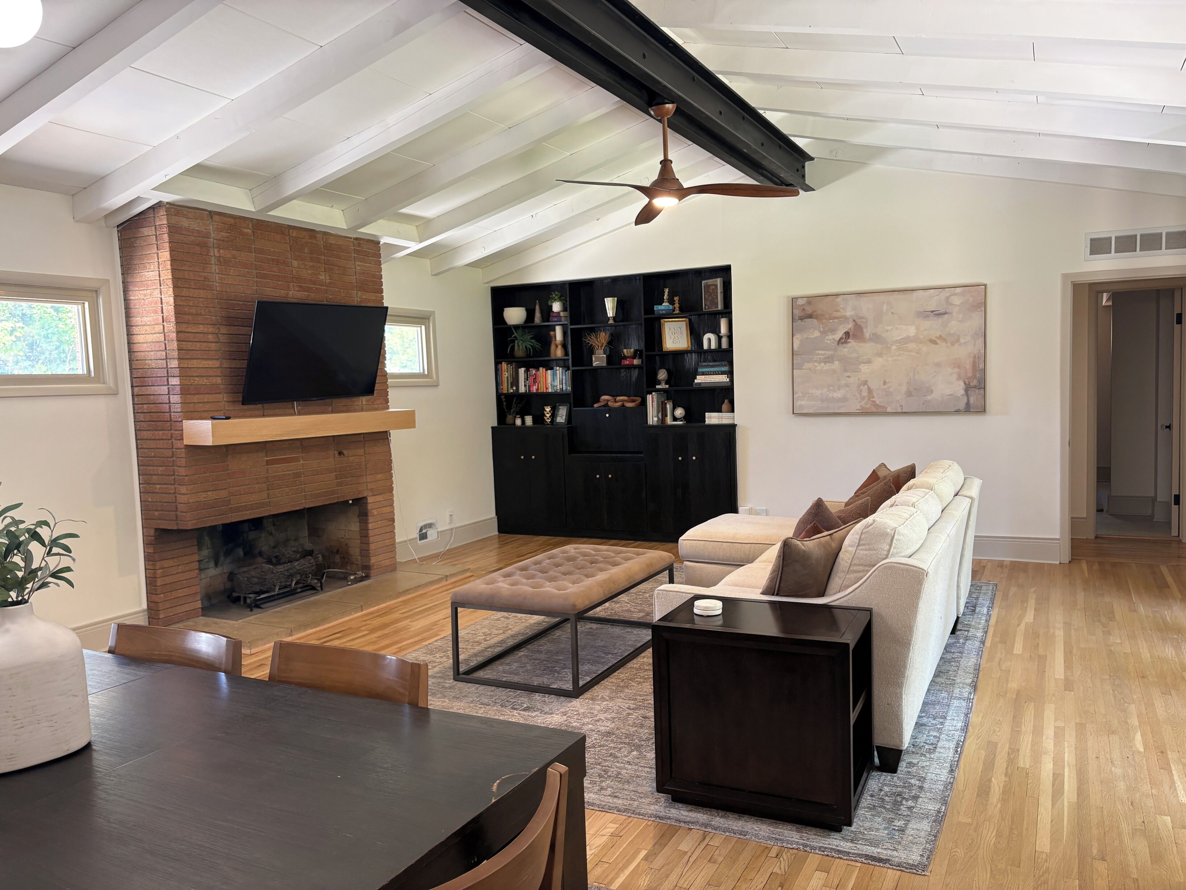 A living room with a brick fireplace, a black entertainment unit, a beige sofa, and a dining area with a table and chairs.
