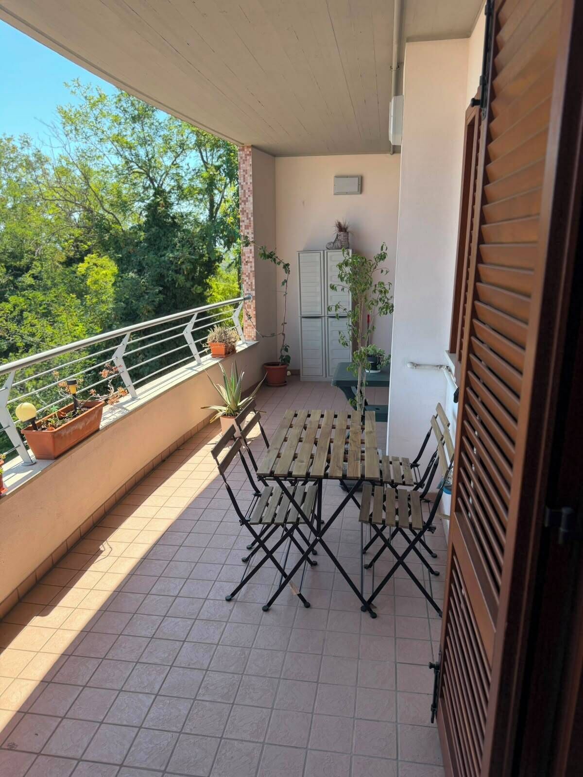 A balcony with a wooden table and chairs, potted plants, and a view of trees.