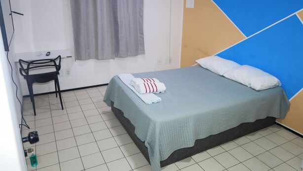 A single bed with a blue headboard, a grey curtain, a small table with a chair, and a tiled floor. - Hostel Way Fortaleza (Fortaleza)