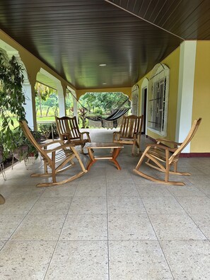 A covered patio area with wooden rocking chairs and a hammock, surrounded by potted plants and a view of greenery.