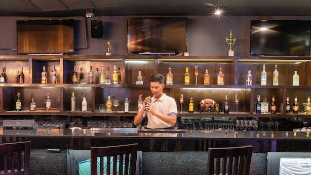 A bar area with multiple shelves of liquor bottles, a bartender preparing drinks, and bar stools.
