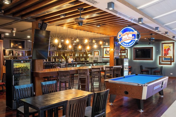 Eine Bar mit einer Poolbillardtisch, Holztischen und -stühlen. Eine Barbediene steht hinter der Theke, neben einem Kühlschrank.