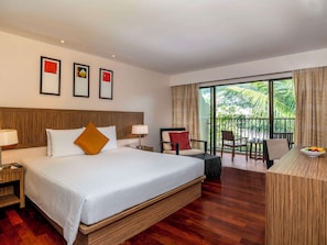 Ein Hotelzimmer mit einem großen Bett, einem Schreibtisch mit Obstschale, einem Sessel und einem Balkon mit Blick auf Palmen.
