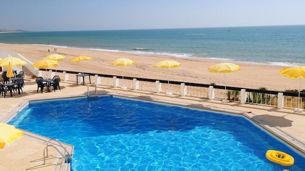 Een zwembad naast een strand, met gele parasols en ligbedden.