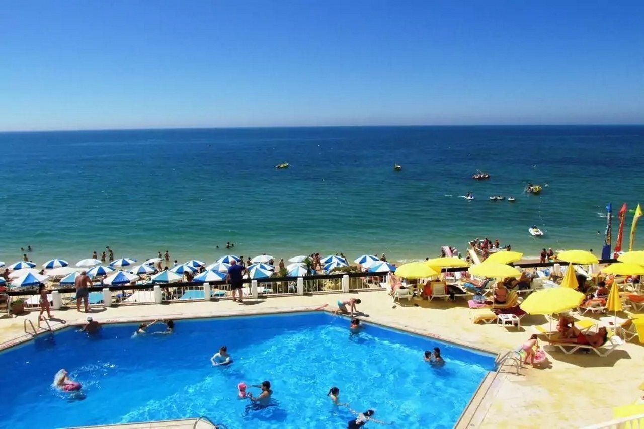 A pool area with people swimming and lounging under umbrellas, overlooking the sea.