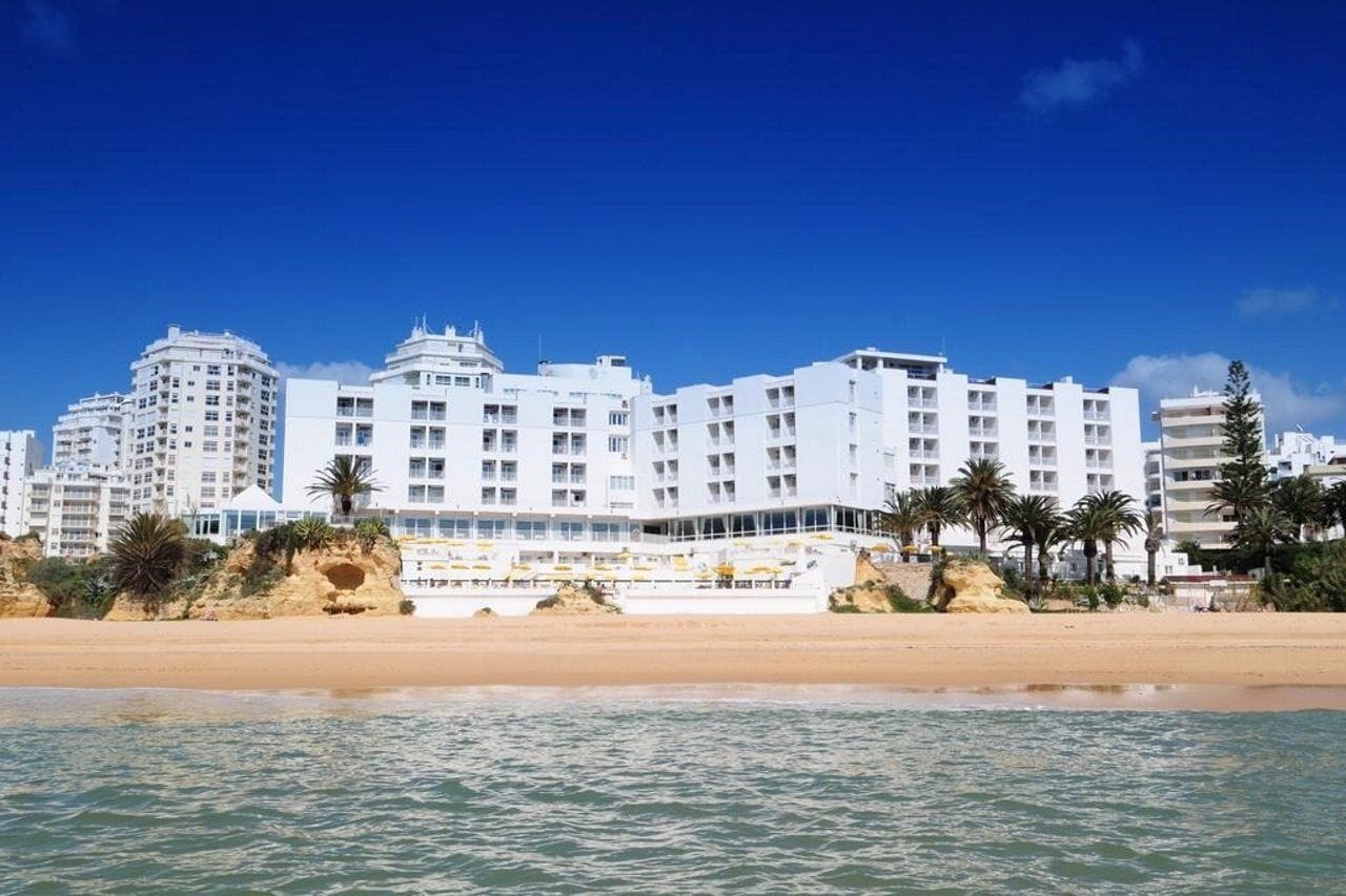 A beachfront hotel with multiple floors, balconies, and a sandy beach in the foreground.