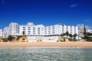 Un hotel frente a la playa con varios pisos, balcones y una playa arenosa en primer plano.