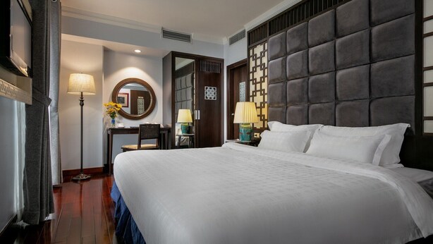 A hotel room with a large bed, a desk with a chair, a lamp, and a wall-mounted mirror. - Grand Minerva Hotel (Hanoi)