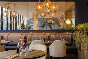 Ein Restaurant mit modernem Ambiente, ausgestattet mit einer langen Sitzbank, runden Tischen und hängenden, kugelförmigen Leuchten.