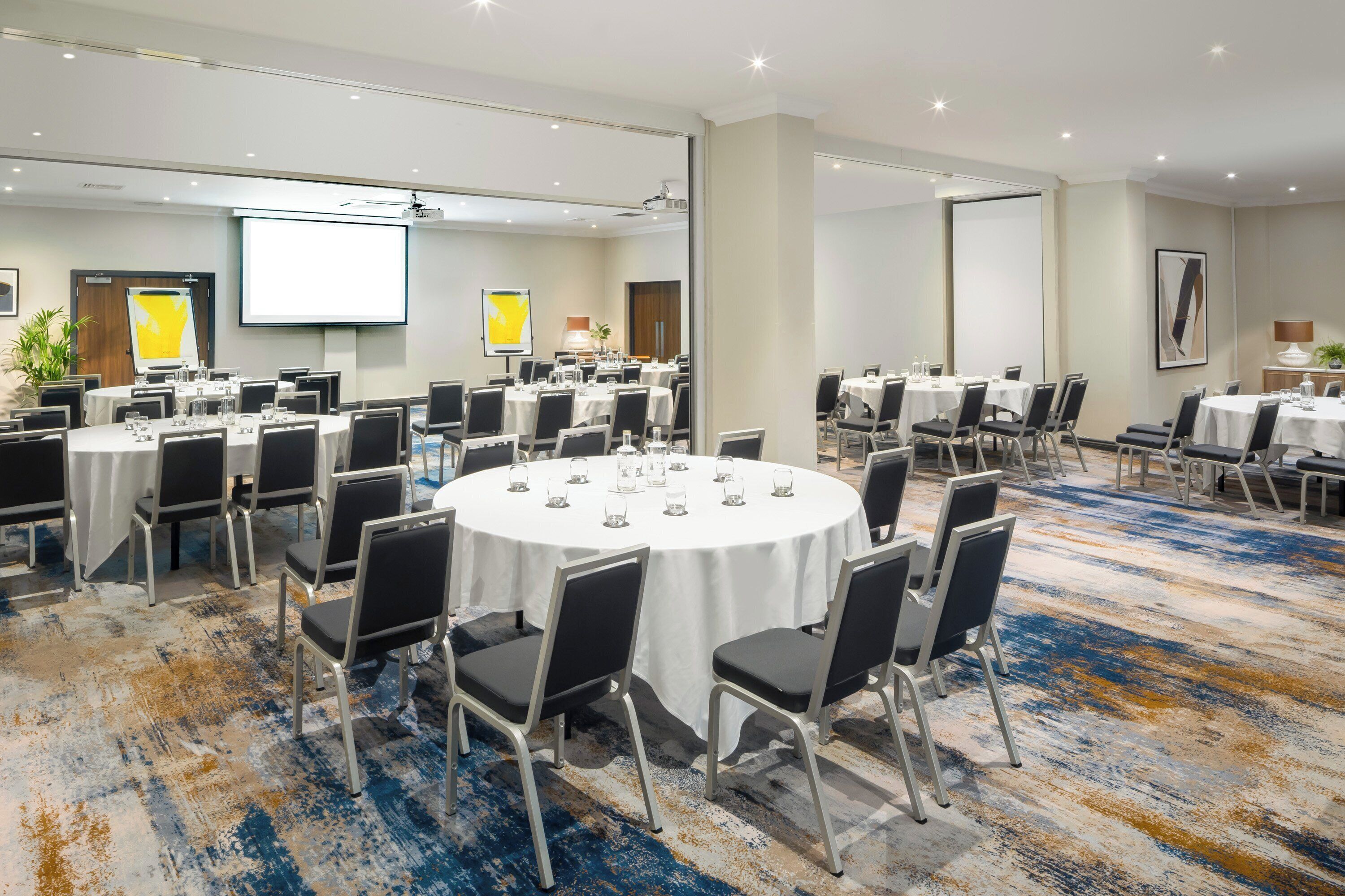 A conference room with multiple round tables, each with chairs, equipped with a projector screen, and a large screen on the wall.