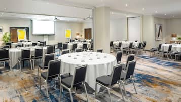 A conference room with multiple round tables, each with chairs, equipped with a projector screen, and a large screen on the wall.