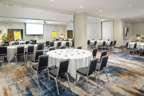 A conference room with multiple round tables, each with chairs, equipped with a projector screen, and a large screen on the wall. - Voco Southampton (Southampton)