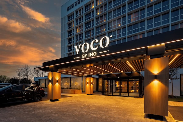 Ein Hotel Eingang mit modernem Design, inklusive eines gut sichtbaren Schildes "VOCO BY IHG" und einem überdachten Bereich.
