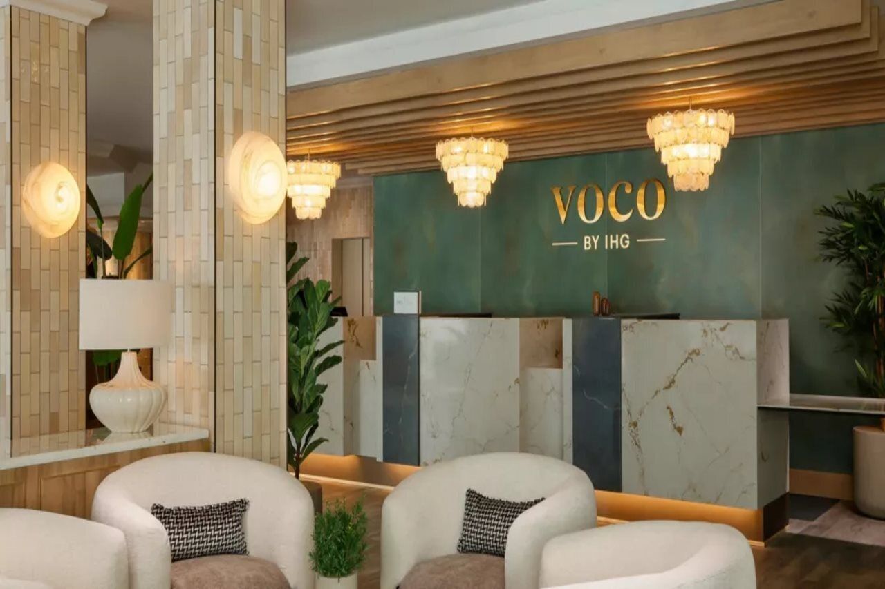 A reception area with a green wall, marble panels, and a sign that reads 'VOCO BY IHG'. There are three chairs with cushions and a potted plant.