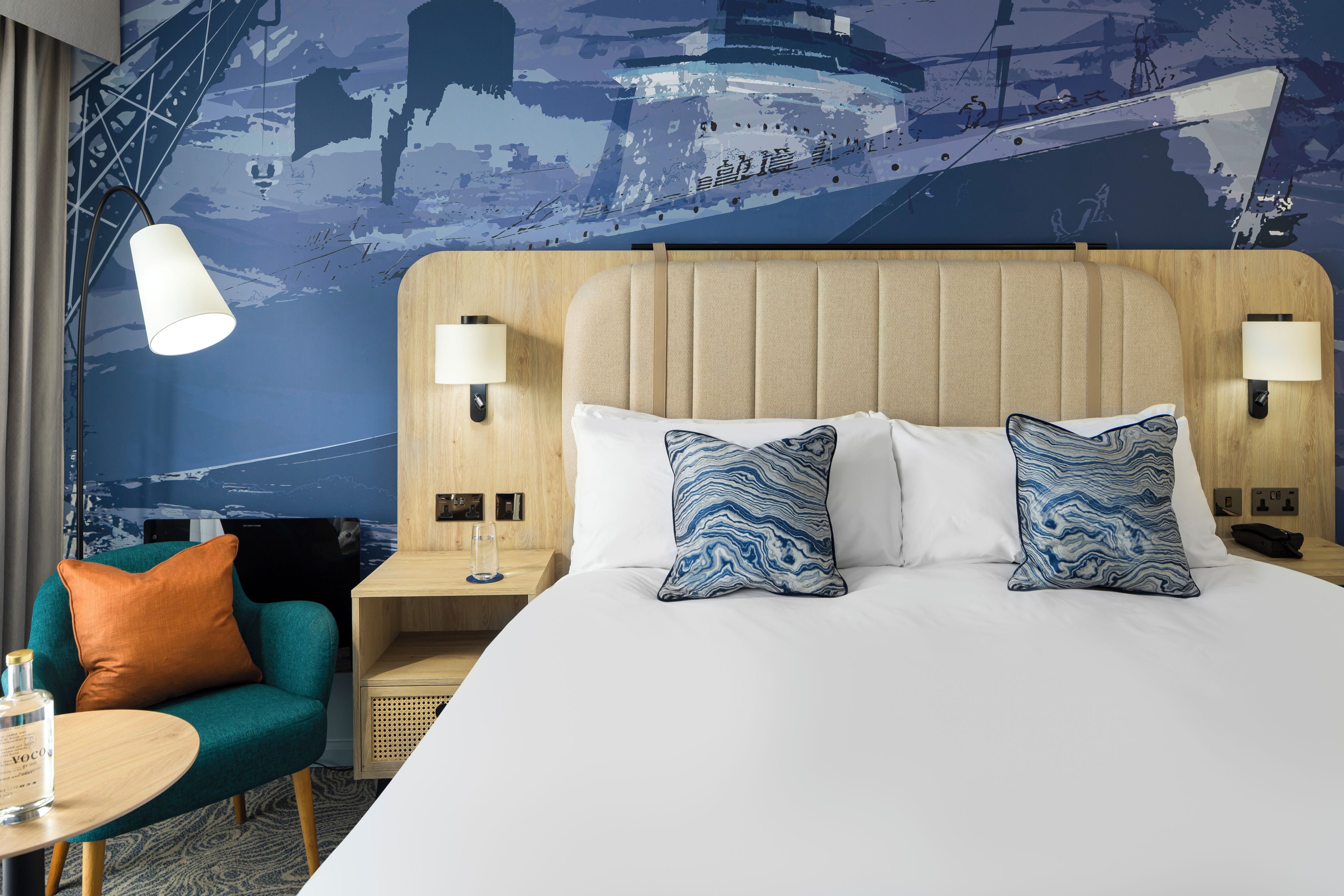 A hotel room with a large bed, two pillows, a nightstand, a lamp, a chair, and a wall mural.