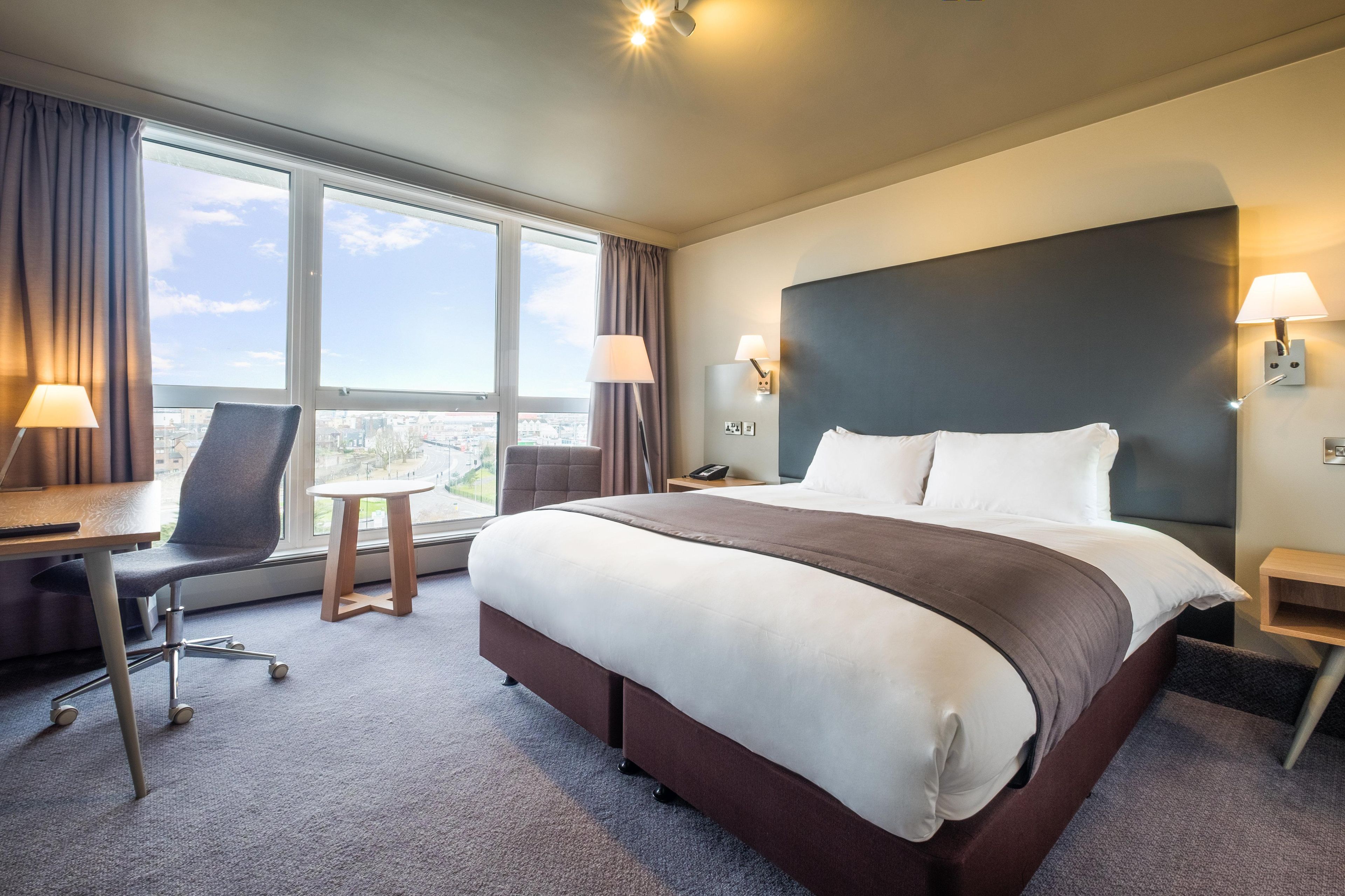 A hotel room with a large bed, a desk, a chair, a small table, and a view of the city through the window.