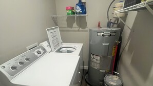 A compact laundry room with a front-loading washer and dryer, a water heater, and a small shelf with cleaning supplies. - 3-bedroom cabin close to Downtown Clayton with a Mountain feel. (Clayton)