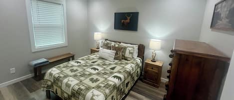 A bedroom with a bed, two bedside tables, a bench, a window with blinds, and a wall-mounted lamp.