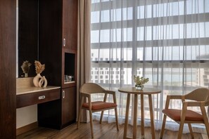 Una habitación con un escritorio de madera, dos sillas, una mesita con un florero y un ventanal con cortinas traslúcidas.