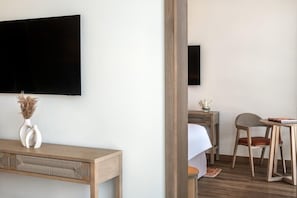 Une chambre d’hôtel dotée d’un téléviseur à écran plat fixé au mur, d’un bureau en bois orné d’un vase de fleurs séchées, d’une petite table avec une chaise et d’un lit recouvert d’un drap blanc.