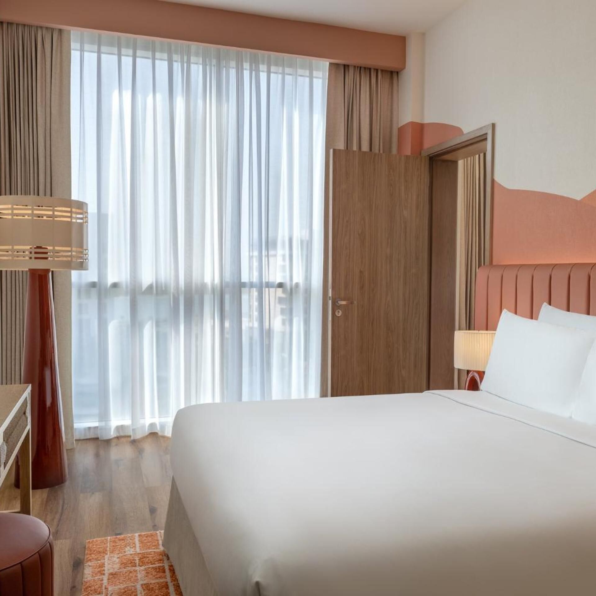 A hotel room with a large bed, a wooden wardrobe, a desk with a lamp, and a view through sheer curtains.