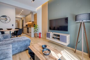 Uma sala de estar moderna com sofá, mesa de centro, televisão de tela plana, luminária de chão e uma área de cozinha com conjunto de mesa e cadeiras.