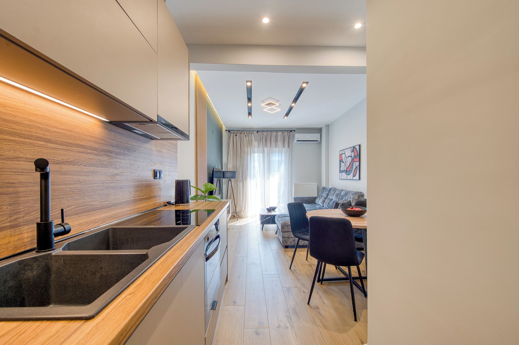 A modern kitchen with a built-in stove, a sink, and a wooden backsplash. There is a dining area with a table and chairs, and a sofa in the background.