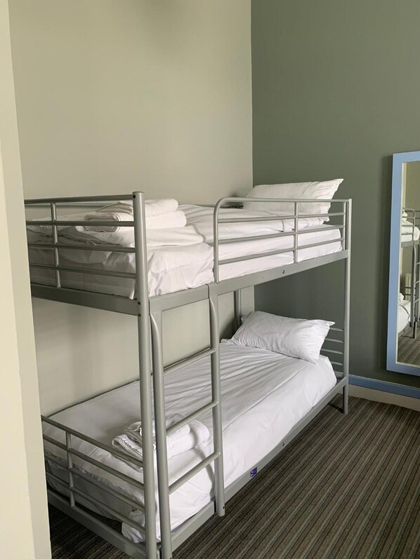Una cama individual con ropa blanca y estructura metálica, en una habitación con piso alfombrado y un espejo en la pared.