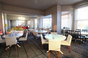 Un comedor de hotel con varias mesas y sillas redondas, una zona de bar y una alfombra estampada.