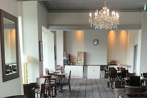 A hotel dining area with a chandelier, a clock, and a kitchen in the background.
