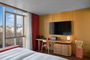 Quarto de hotel com cama, escrivaninha com televisão de tela plana, cadeira, telefone vermelho, pequeno alto-falante e vaso com plantas secas.