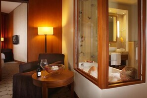 Habitación de hotel con pared de madera, una mesa redonda con una botella de vino y una copa, un ventanal con vista a un baño, y un cuadro en la pared.