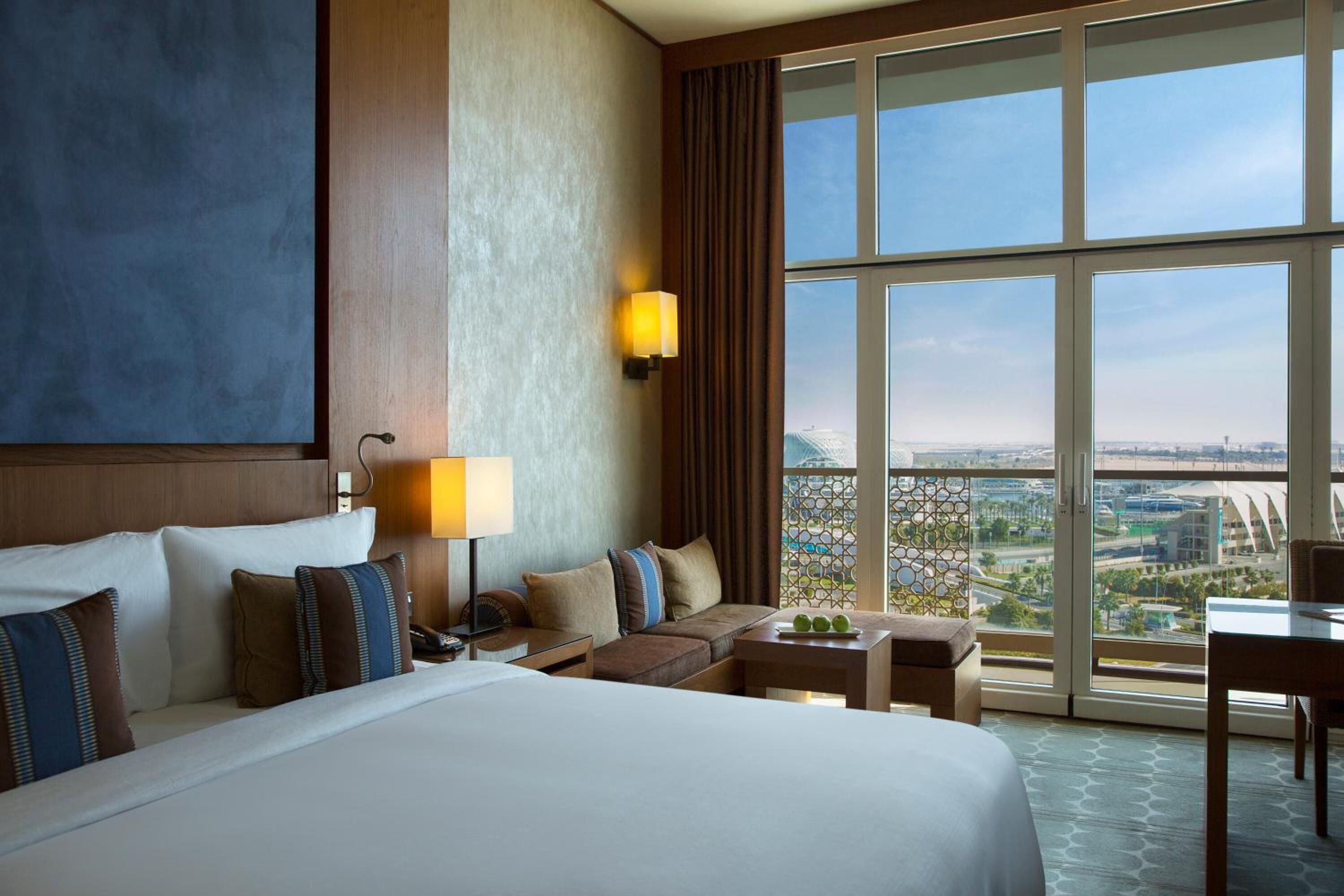 A hotel room with a large bed, a desk, a chair, a sofa, and a view of the city through large windows.