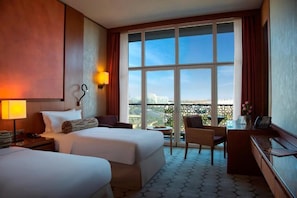 Quarto de hotel com duas camas, uma escrivaninha, uma cadeira, uma janela grande com vista e chão estampado.