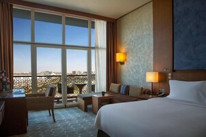 Habitación de hotel con una cama grande, un escritorio, una silla, una mesita y vistas a la ciudad a través de amplios ventanales.