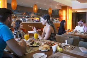 Uma área de refeições com mesas postas, um bar ao fundo e iluminação pendente laranja.