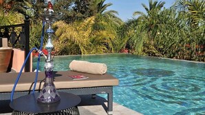 Een terras bij het zwembad met een shisha op een tafel, een opgerolde handdoek, en een helder zwembad omgeven door tropische beplanting.